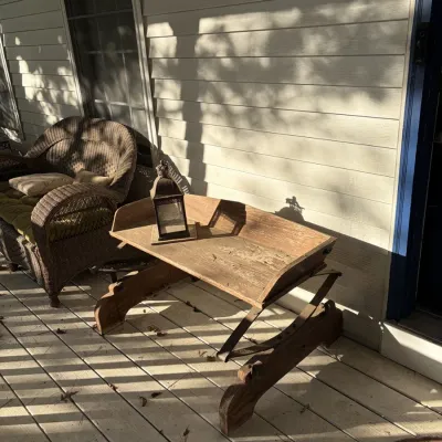 Sale Photo Thumbnail #110: Rustic wooden trough-style table with iron accent straps, paired with a small metal lantern on a porch next to a wicker armchair.