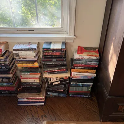 Sale Photo Thumbnail #60: Several neat stacks of contemporary fiction and non-fiction paperback and hardcover books arranged on a hardwood floor under a window.