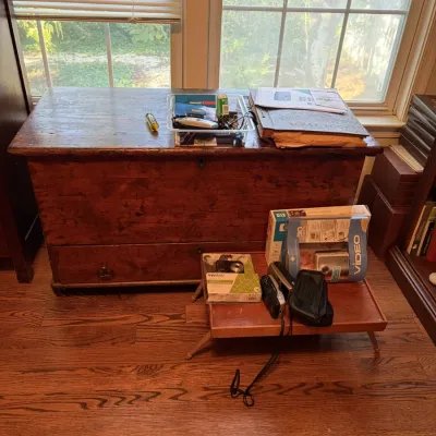 Sale Photo Thumbnail #92: Rustic antique wooden blanket chest featuring a single bottom drawer, alongside a small folding bed tray holding various electronics and cameras.