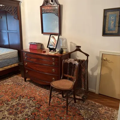 Sale Photo Thumbnail #128: Traditional mahogany bow-front dresser paired with a matching broken-pediment wall mirror, a cane-seat side chair, and an intricate Persian-style area rug.