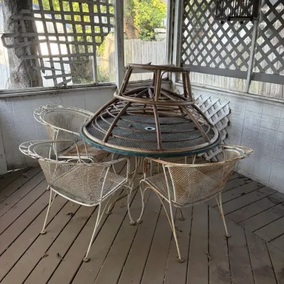 Sale Photo Thumbnail #103: Vintage wrought iron patio set with a mesh top table and four matching chairs, showing rust and wear. A rattan or bamboo base structure sits stacked upside down on top of the table.