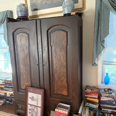 Sale Photo Thumbnail #68: Large antique dark wood armoire with burl wood paneling on the doors, flanked by stacks of books and decorative porcelain pieces.