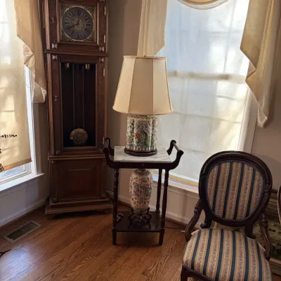 Sale Photo Thumbnail #14: Traditional wooden grandfather clock standing in a room corner beside a marble-top accent table with an Asian-style ceramic lamp and a blue-and-gold striped upholstered side chair.