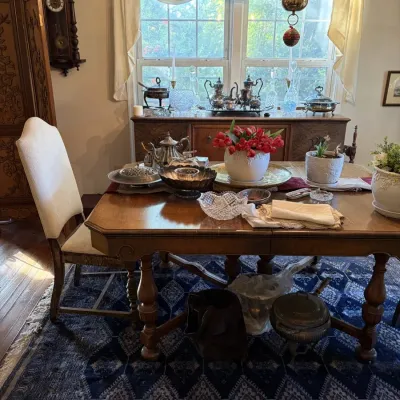 Sale Photo Thumbnail #106: Traditional wooden dining table and upholstered chair set with silver serving pieces, crystal glassware, a wall clock, and a matching sideboard in the background.