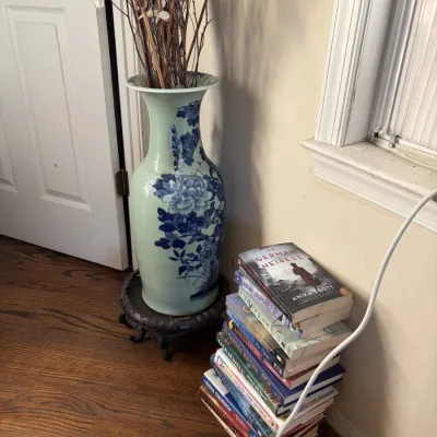 Sale Photo Thumbnail #59: Large blue and white floral ceramic floor vase on a carved wooden base, displayed alongside a stack of contemporary paperback and hardcover books.