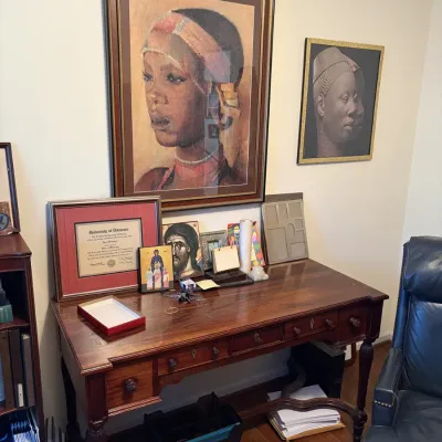 Sale Photo Thumbnail #74: Traditional mahogany writing desk with turned legs and drawers, surrounded by framed artwork including African portraits, a University of Delaware diploma, and religious icons.