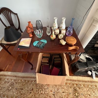 Sale Photo Thumbnail #24: Wooden side table displaying an array of glassware, ceramic vases, and decorative heart-shaped stained glass pieces, with a box of vintage books stored underneath.