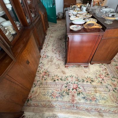 Sale Photo Thumbnail #39: Large pastel floral Aubusson-style wool area rug with fringed ends, displayed among traditional dark wood china cabinets and dressers loaded with vintage dishware.