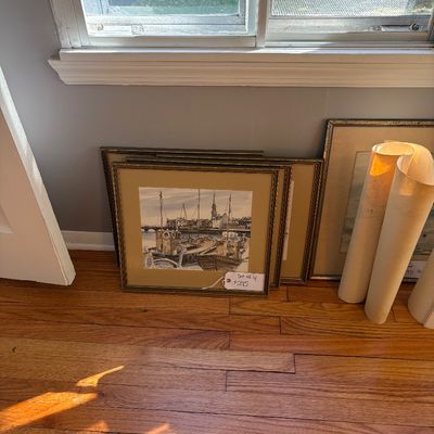 Sale Photo Thumbnail #3: Set of four framed harbor scene artwork prints resting against a wall on hardwood flooring, alongside rolled posters.