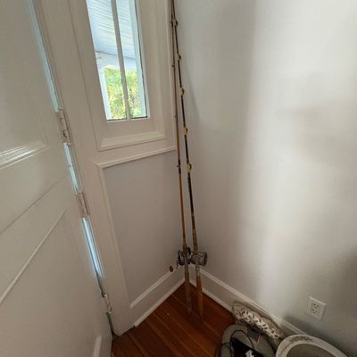 Sale Photo Thumbnail #33: Pair of vintage saltwater fishing rods with attached conventional reels leaning against a corner wall.