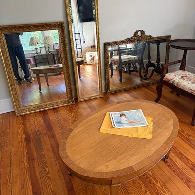 Sale Photo Thumbnail #17: An oval wood coffee table with banded inlay, displayed alongside three ornate gilt-framed wall mirrors and an upholstered accent chair.
