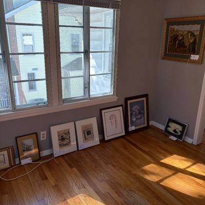 Sale Photo Thumbnail #6: Collection of framed artwork, prints, and drawings displayed on the floor and wall of a room with hardwood flooring.