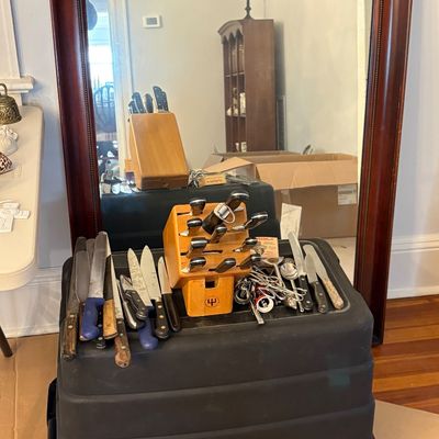 Sale Photo Thumbnail #42: Collection of assorted kitchen knives, cutlery, and a wooden knife block set displayed on a plastic tote in front of a large mahogany-framed mirror.