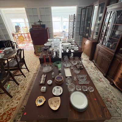 Sale Photo Thumbnail #34: A wide variety of vintage glassware, china, porcelain dishes, and trinkets displayed on long wooden tables alongside traditional china cabinets.