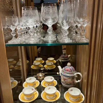 Display cabinet filled with fine crystal stemware, a floral teapot, and yellow porcelain teacups with saucers.