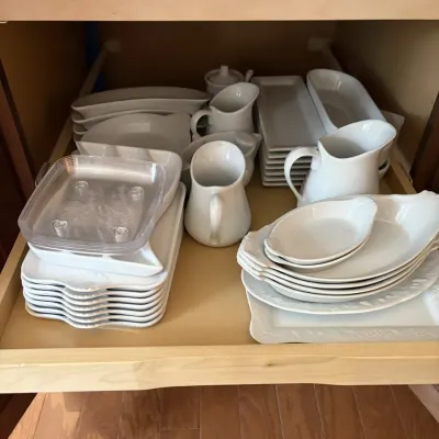 Pull-out cabinet drawer filled with white ceramic serving pieces, including creamers, platters, rectangular dishes, and small bowls.