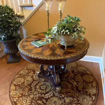 Traditional round foyer table with marquetry wood inlay and ornate gilded trim, displayed on a matching floral area rug with decorative...