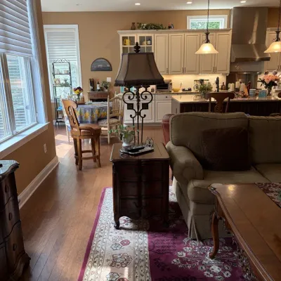 Open-concept living space furnished with a tan upholstered sofa, dark wood end table with a tall decorative lamp, traditional patterned...