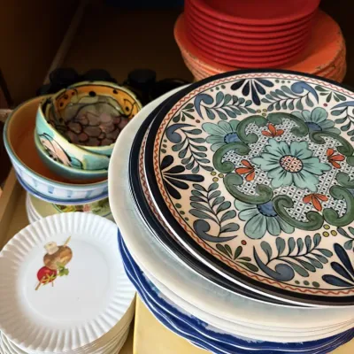 Collection of assorted dinnerware inside a cabinet, featuring Talavera-style hand-painted ceramic plates and bowls, white fruit-patterned...