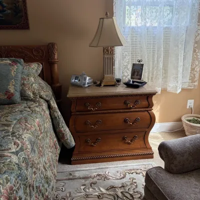 Traditional Bombe nightstand with ornate bronze hardware and marble top, flanked by a bed and an armchair in a bedroom setting.
