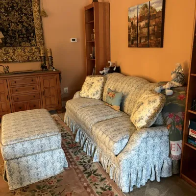 Traditional skirted sofa and matching ottoman in a damask pattern, paired with a floral area rug, wooden sideboard, and wall art.