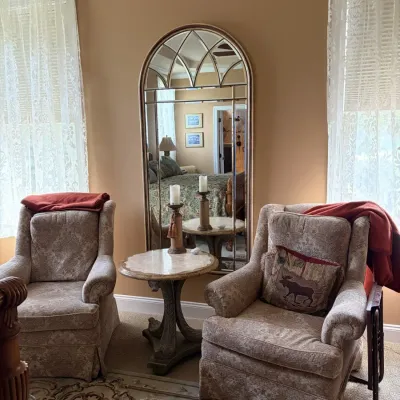 Pair of beige patterned upholstered armchairs flanking a round pedestal side table, with a large cathedral-style arch floor mirror set...