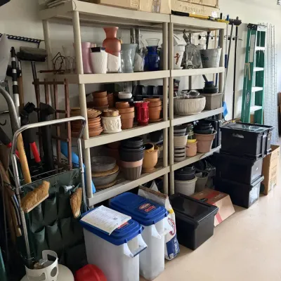 Garage storage setup filled with gardening supplies, including terracotta planters, plastic pots, pet food containers, lawn tools, and a...