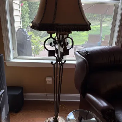 Ornate wrought iron floor lamp with a heavy carved pedestal base and a bell lampshade featuring tassel fringe detail.