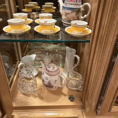 Collection of fine porcelain and glass tea service items displayed inside a mirrored wooden hutch, featuring yellow-banded teacups with...