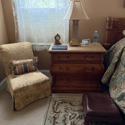 Traditional bedroom furnishings featuring a three-drawer bombé nightstand with a stone top, a pillar-style table lamp, and an armless...
