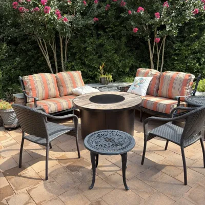 Outdoor patio furniture set featuring a round propane fire pit table, two cushioned metal loveseats with orange striped cushions, and...