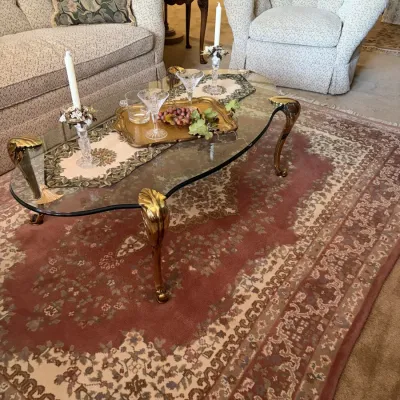 Vintage glass-top coffee table with brass legs, set on a rose-and-cream patterned area rug in a traditional living room.