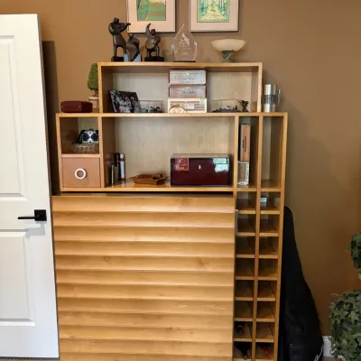 Contemporary light wood bar cabinet featuring a louvered front door, side wine bottle storage rack, and multi-tier upper display shelving...