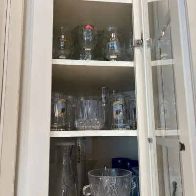 White kitchen cabinet filled with various glassware, including branded beer goblets, glass mugs, carafes, and a cut glass ice bucket.