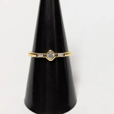 Yellow gold ring featuring a central bezel-set princess cut clear stone flanked by two baguette accent stones, displayed on a black ring...