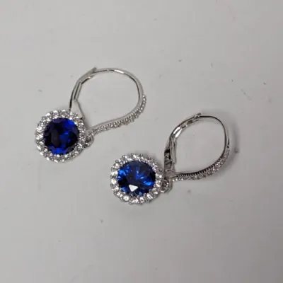 Pair of sterling silver drop earrings featuring round-cut synthetic blue sapphire stones surrounded by clear cubic zirconia halos with...