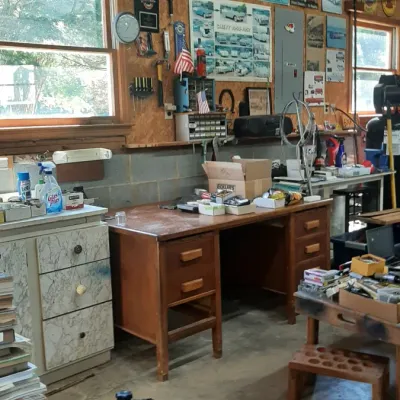 Workshop and garage area featuring a push lawn mower, vintage wooden desk, cabinets, tool storage, and automotive memorabilia on OSB walls.
