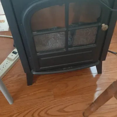 Compact black electric fireplace heater featuring a traditional stove design with a glass panel door and fake log display.