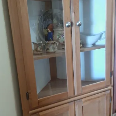 Tall light wood corner display cabinet with glass-front upper doors, lower cabinet doors, and silver-tone knobs.