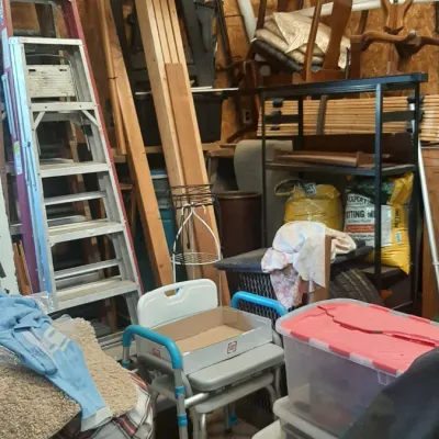 Workshop and storage area filled with stepladders, plastic storage totes, lumber, and stacked dining chairs.