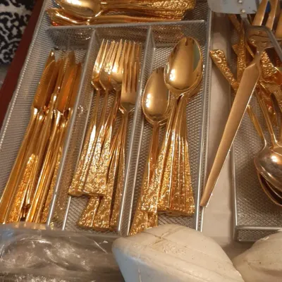 Gold-toned flatware set with textured handles arranged in wire organizer trays, accompanied by serving utensils.