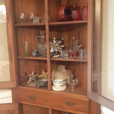 Vintage early American-style pine corner hutch with glass-paned doors and a decorative broken arch pediment, filled with various...