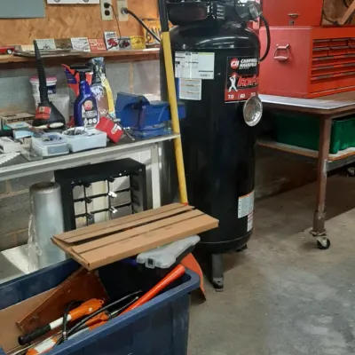 A Campbell Hausfeld Iron Force upright air compressor standing in a workshop alongside a red rolling tool chest, plastic storage totes...
