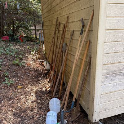 Sale Photo Thumbnail #71: Collection of assortment long-handled yard tools leaning against a shed, including shovels, rakes, and a scythe.