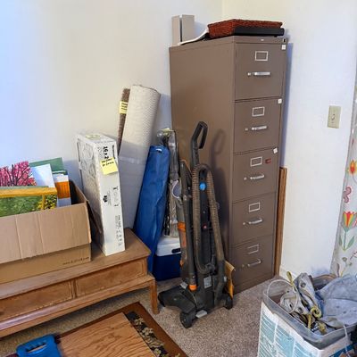 Sale Photo Thumbnail #7: Tall brown metal five-drawer filing cabinet shown alongside an upright vacuum cleaner, rolled area rug, boxed air filters, and various household items.