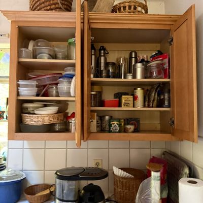 Sale Photo Thumbnail #33: Open wooden kitchen cabinets filled with plastic storage containers, travel mugs, and tea tins above a countertop with various small kitchen appliances and woven baskets.