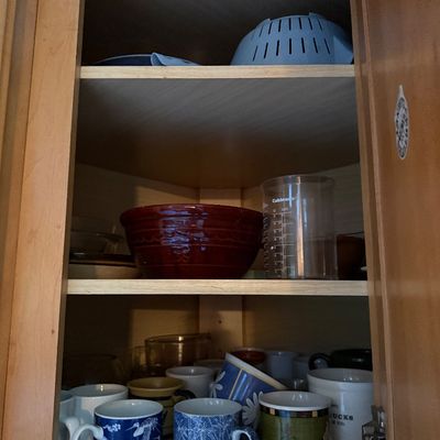 Sale Photo Thumbnail #35: Kitchen cabinet filled with assorted ceramic coffee mugs, a red glazed stoneware mixing bowl, a Cuisinart measuring cup, and plastic kitchenware.