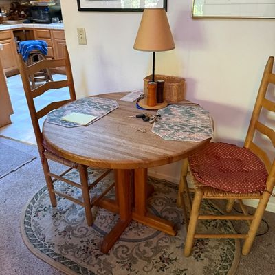 Sale Photo Thumbnail #57: Round wooden pedestal dining table paired with two ladder-back chairs featuring rush seats and red seat cushions, set over an oval patterned area rug.