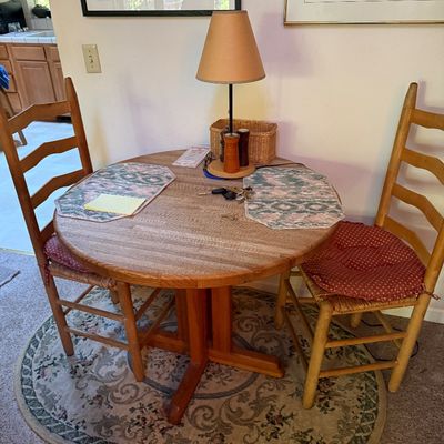 Sale Photo Thumbnail #22: Round wooden butcher-block style pedestal table paired with two ladder-back dining chairs with woven seats and red cushions.