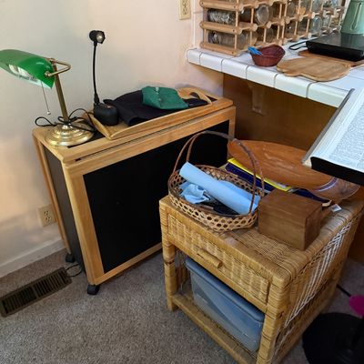 Sale Photo Thumbnail #24: Natural wicker side table paired with a rolling wooden cabinet with a black front, topped with a green banker's lamp and small household items.